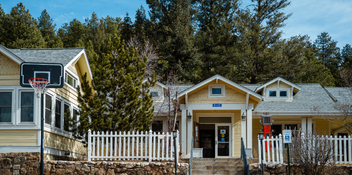 Exterior photo of the Evergreen Resiliency Center, a building with pale yellow wood siding with evergreen trees against a clear blue sky in the background and a portable basketball hoop on the street in the foreground. 